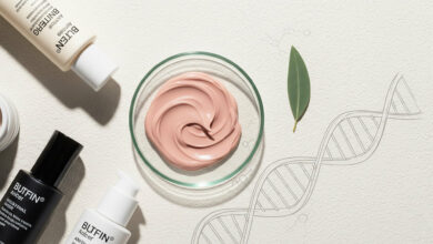 A flat-lay of anti-aging skincare products, including cream and charcoal bottles, a petri dish with dusty rose-colored cream, and a sage green leaf, arranged on a cream background with a DNA helix illustration.