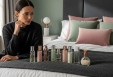 Woman contemplating a collection of unbranded body mists in a chic bedroom with dusty rose, sage green, and charcoal decor, for fragrance and beauty product reviews.