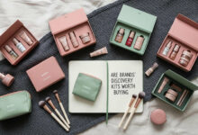 A flat lay photograph of various beauty discovery kits. The kits are in dusty rose and sage green boxes and pouches, arranged on a cream and charcoal textured fabric. In the middle, an open notebook reads, "ARE BRANDS' DISCOVERY KITS WORTH BUYING," surrounded by small makeup brushes and miniature beauty products.