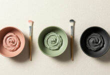 image of three clay masks in a horizontal row on a cream-colored textured surface. From left to right: a dusty rose clay mask in a matching bowl with a brush, a sage green clay mask in a matching bowl with a brush, and a charcoal clay mask in a black bowl with a brush. Each mask has a visible swirl, indicating its creamy texture.