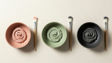 image of three clay masks in a horizontal row on a cream-colored textured surface. From left to right: a dusty rose clay mask in a matching bowl with a brush, a sage green clay mask in a matching bowl with a brush, and a charcoal clay mask in a black bowl with a brush. Each mask has a visible swirl, indicating its creamy texture.