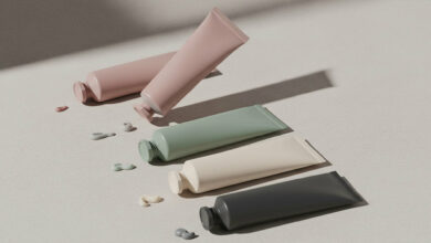 A photorealistic image from a high angle, displaying four tubes of hand cream in dusty rose, sage green, cream, and charcoal, ranked in a diagonal line. A small amount of matching cream is squeezed out next to each tube on the textured, neutral-colored background, all under soft, natural lighting.
