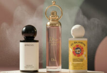 A photorealistic image showcasing three popular hair mists, each standing on individual cream-colored stone pedestals against an abstract background of dusty rose and sage green geometric shapes. From left to right: a bottle of Byredo Hair Perfume, a pink bottle of Dior J'adore Hair Mist with a golden ring top, and a yellow bottle of Sol de Janeiro Cheirosa '62 Hair & Body Mist. A fine mist emanates around the bottles, conveying the idea of fragrance.