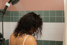 A photorealistic image of a person with wet, curly hair viewed from behind in a shower. Their head is slightly bowed as water from a filtered, charcoal-colored showerhead sprays onto their hair. The bathroom features sage green and white tiles, a dusty rose wall, and a cream shower curtain, creating a stylish backdrop for the scene of hair care frustration.