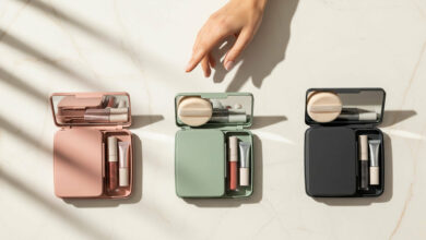 image of three open makeup touch-up kits in dusty rose, sage green, and charcoal, arranged in a row on a cream-colored marble surface. Each kit contains a powder puff, a lip gloss, and other small items. A woman's hand is seen hovering over the kits, as if thoughtfully choosing one.