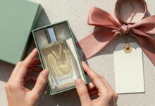 A complete buying guide on how to choose perfume as a gift, shown as a flat-lay of hands placing a perfume bottle into a sage green gift box with a dusty rose ribbon and tag.