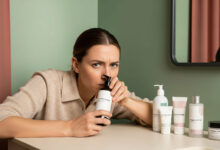 mage of a woman in a cream-colored sweater, leaning over a bathroom counter and suspiciously sniffing a bottle of "fragrance-free" product. Her brow is furrowed in concentration. On the counter, there is a lineup of other minimalist, unbranded "fragrance-free" products. The bathroom has a modern aesthetic with a sage green wall, a charcoal-framed mirror, and a dusty rose shower curtain in the background.