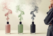 Woman contemplates fragrance sillage from three perfume bottles (dusty rose, sage green, charcoal) emitting colored smoke, representing scent concentration and projection.