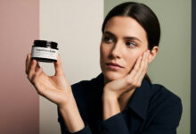 A photorealistic image of a woman in a charcoal shirt holding a jar of Augustinus Bader cream. She has a thoughtful and questioning expression, resting her cheek on one hand while closely inspecting the jar. The background is composed of modern color blocks in dusty rose, sage green, and cream.