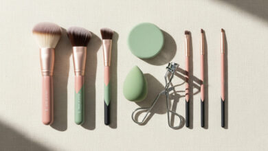 A beginner's guide to makeup brushes, sponges, and tools, illustrated by a flat-lay of various brushes with dusty rose and sage green handles, a green sponge, and an eyelash curler neatly arranged on a cream background.