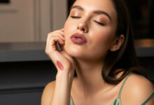 A close-up, photorealistic image of a woman with her eyes closed in a moment of tranquil reflection. She rests her chin on her hand, which has a swatch of the same dusty rose matte lipstick she is wearing. Her sage green camisole strap is slightly off her shoulder, suggesting a long night out. The background is a soft-focus blend of cream and charcoal.