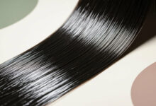 A complete scientific guide to getting shiny, glossy hair, illustrated by an extreme close-up of a perfectly smooth and reflective lock of dark hair against a minimalist, colored background.