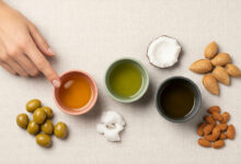 showcasing natural oils for nail health: olive oil, coconut oil, and almond oil. Three small bowls, colored dusty rose, sage green, and charcoal, contain the respective oils. A woman's hand points to the olive oil. The scene is enriched with corresponding raw ingredients: green olives, a cracked coconut, whole almonds, and shelled almonds, all arranged on a cream-colored textured surface for enhanced visual appeal and search ranking for natural beauty and nail care topics.