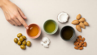 showcasing natural oils for nail health: olive oil, coconut oil, and almond oil. Three small bowls, colored dusty rose, sage green, and charcoal, contain the respective oils. A woman's hand points to the olive oil. The scene is enriched with corresponding raw ingredients: green olives, a cracked coconut, whole almonds, and shelled almonds, all arranged on a cream-colored textured surface for enhanced visual appeal and search ranking for natural beauty and nail care topics.