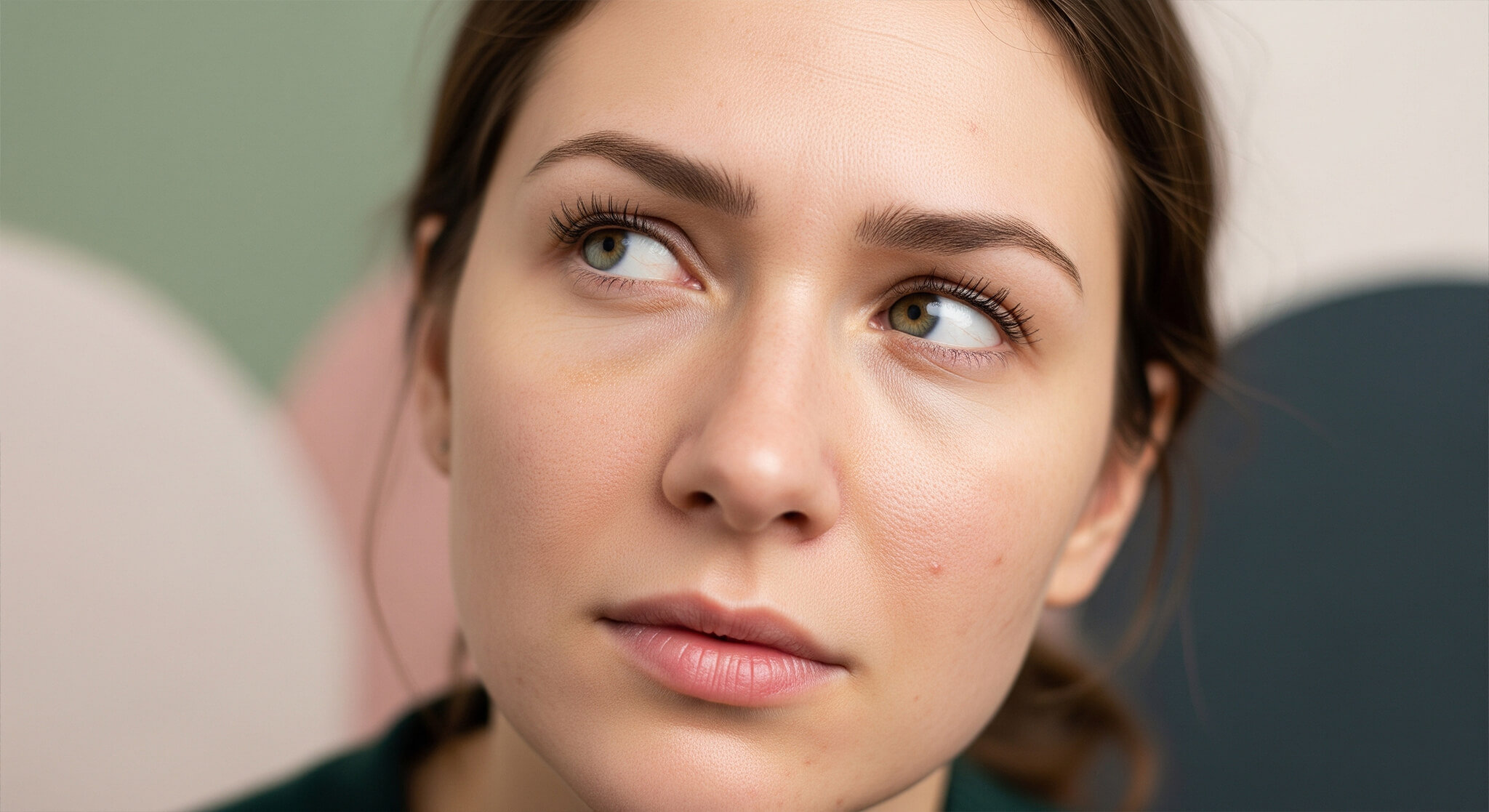 A close-up, photorealistic image of a woman's face. She has a pensive expression, with her eyes looking up and to the side. Her skin is shown in realistic detail. The background is a soft-focus, abstract blend of dusty rose, sage green, cream, and charcoal.