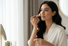 A gorgeous woman in a white robe applies foundation with a brush at a vanity, surrounded by various foundation sticks, for a review of the best foundation sticks for quick application.