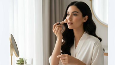 A gorgeous woman in a white robe applies foundation with a brush at a vanity, surrounded by various foundation sticks, for a review of the best foundation sticks for quick application.