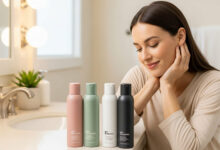Smiling woman examining four dry shampoo bottles (dusty rose, sage green, cream, charcoal) in a clean bathroom, highlighting gentle formulas for sensitive scalps and hair care reviews.