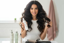 A gorgeous woman with voluminous wavy hair holds up a bottle of heat protectant, surrounded by other hair products and a curling iron, reviewed as the best heat protectants that maintain hair volume.