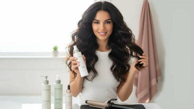 A gorgeous woman with voluminous wavy hair holds up a bottle of heat protectant, surrounded by other hair products and a curling iron, reviewed as the best heat protectants that maintain hair volume.
