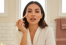 A gorgeous woman applies peach color corrector under her eye with a brush, surrounded by various peach and orange color correctors, for an article on neutralizing severe dark circles.