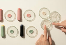 Hands testing lip balm ingredients: a flat lay of five lip balm tubes (dusty rose, sage green, charcoal) and corresponding swatches/petroleum jelly in petri dishes, for ingredient analysis and beauty product comparisons.
