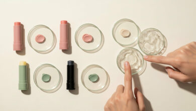 Hands testing lip balm ingredients: a flat lay of five lip balm tubes (dusty rose, sage green, charcoal) and corresponding swatches/petroleum jelly in petri dishes, for ingredient analysis and beauty product comparisons.