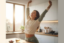 A gorgeous, energetic woman in a bright kitchen stretches with a happy smile, next to a cup of tea and a journal, illustrating increased energy levels from intermittent fasting.