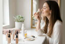 A woman applies foundation at a vanity, with face oils and foundation bottles around, illustrating how mixing face oil with foundation can help achieve a dewier base.