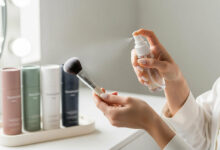 Woman cleaning a makeup brush with sanitizing spray, with various colorful bottles on a vanity, for a review of the best brush cleaners.