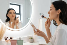 Woman in a bright, modern bathroom vanity area, looking into a round illuminated mirror while holding CC cream and a brush, contemplating her makeup choice. Two generic cosmetic bottles labeled "CC Cream" and "Foundation" are visible on the vanity.