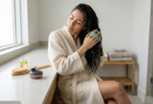 a woman in a bright bathroom environment testing various scalp exfoliation and massage brushes, arranged on a counter amidst brand-colored spa accessories.