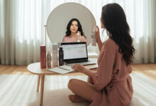 a woman with dark hair in a bright room, applying dry shampoo and checking her hair in a mirror for any white cast, as part of a 'Tested by Sylvaia' review