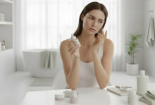 A thoughtful woman with slightly dehydrated skin examines a serum bottle, touching her cheek in a bright, modern bathroom.