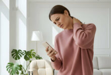 A woman holds her neck in discomfort, looking at her smartphone in a bright, modern living room.