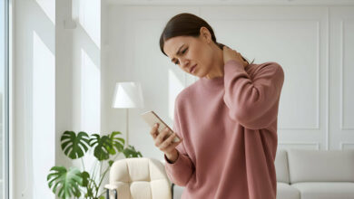 A woman holds her neck in discomfort, looking at her smartphone in a bright, modern living room.