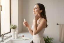 A serene woman in her late 20s, Mia, applies serum to her clear, radiant skin in a bright, modern bathroom. Minimalist products and soft natural light create a calm atmosphere, suggesting confidence gained from a consistent self-care routine.