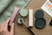 flat lay captures a hand reaching for a dusty rose eye primer amidst a curated selection of sage green, cream, and charcoal makeup products arranged on a rustic wooden surface