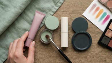 flat lay captures a hand reaching for a dusty rose eye primer amidst a curated selection of sage green, cream, and charcoal makeup products arranged on a rustic wooden surface