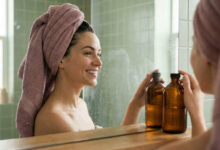 a gorgeous woman with a dusty rose towel wrapped around her hair, smiling happily at her reflection in a steamy, sage-green tiled bathroom as she touches an amber glass bottle.