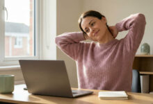 a gorgeous woman in a dusty rose sweater experiencing relief as she stretches her neck at a sunlit desk, accompanied by a sage green mug.