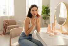 A radiant young woman with a knowing smile holds a K-Beauty lip product in a bright, modern room.