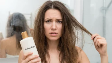 A distressed woman with dry, frizzy hair holding a bottle of 'Sulfate-Free' shampoo in a bathroom