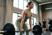 A determined woman in sage green activewear performing a barbell deadlift in a modern, industrial-style gym.