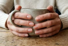 A close-up photograph of a woman's hands with strong, well-cared-for natural nails, lightly coated with clear polish, holding a ceramic mug on a wooden table. This illustrates the concept of healthy nails as a daily accessory