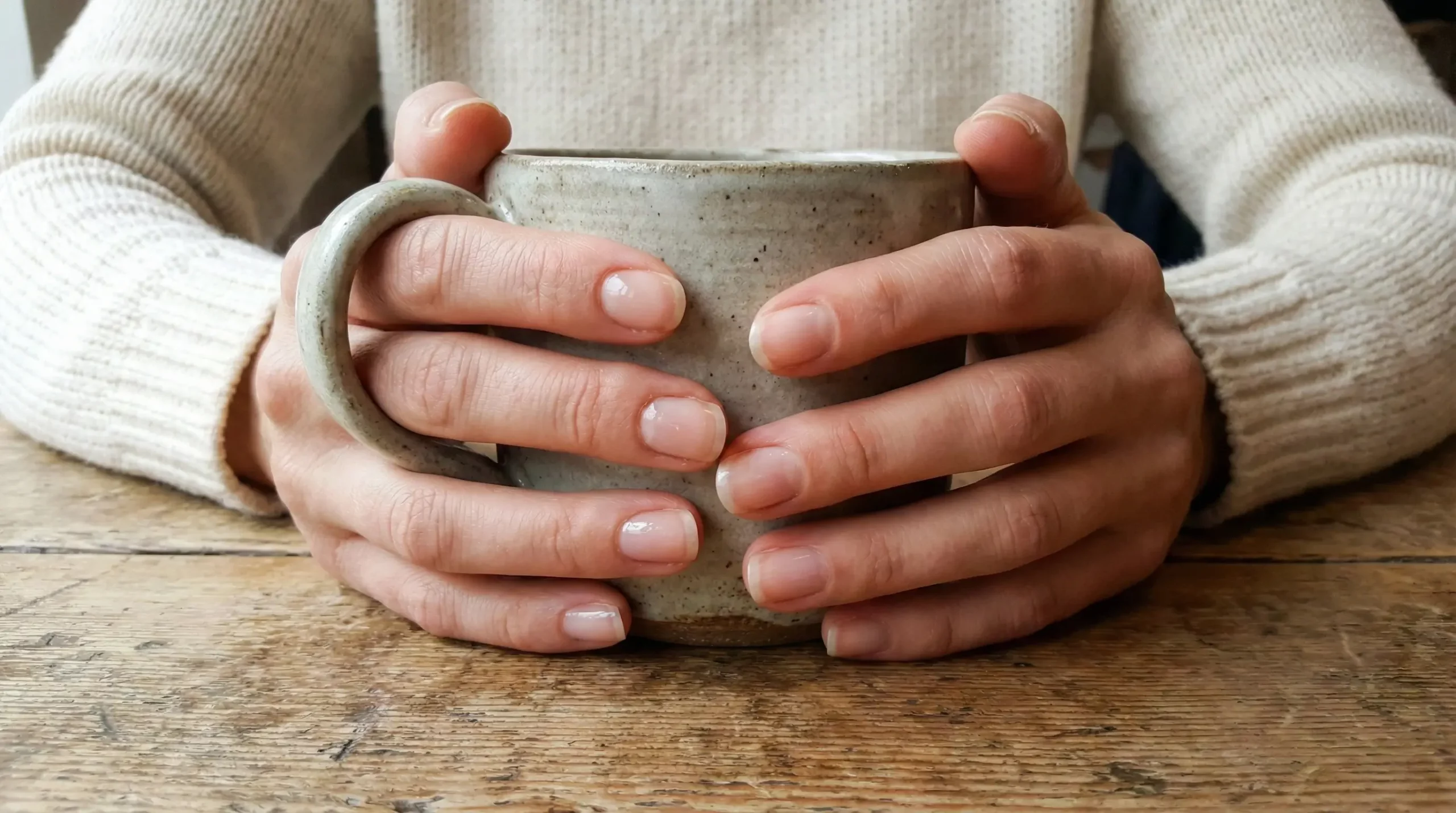 A close-up photograph of a woman's hands with strong, well-cared-for natural nails, lightly coated with clear polish, holding a ceramic mug on a wooden table. This illustrates the concept of healthy nails as a daily accessory