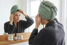 A woman wearing a towel turban examines her scalp in a bathroom mirror, looking for signs of itch, flakes, or oil, with specific scalp care products visible on the counter