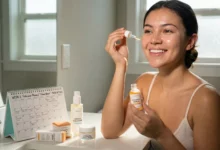 A smiling woman with glowing skin applying Vitamin C serum during her follicular phase 'Glow Week', alongside a calendar and other gentle skincare products in a sunny bathroom.