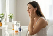 A woman in her late 30s applies an anti-aging serum to her face as part of a skincare routine at a bright vanity, with products including SPF 50 and retinol cream visible.