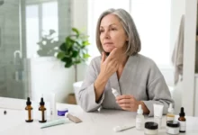 Mature woman with gray hair in a bathrobe examining a blemish on her chin in a bright bathroom, with various skincare products on the vanity.